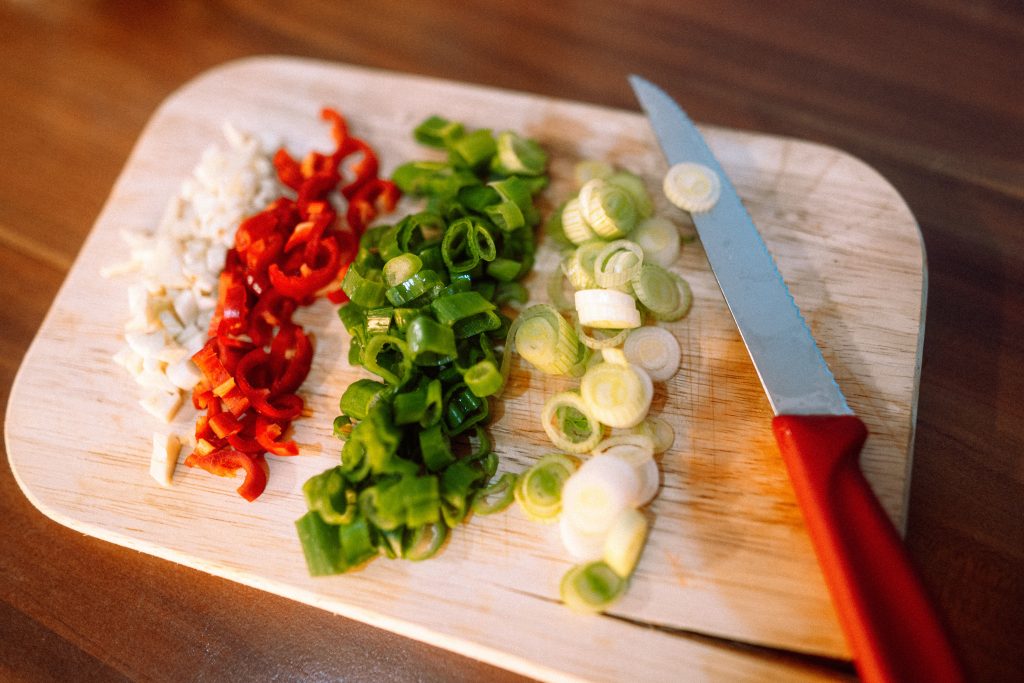 Image shows chopping board, full of chopped vegetables including red peppers, green spring onions and leeks, and a large chefs knife