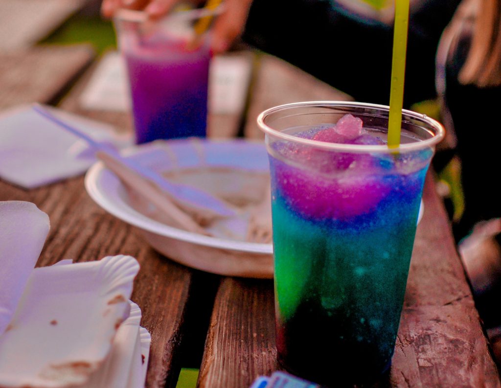 Image shows plastic cup containing very colourful, iced slushy drink with a yellow straw sitting on a wooden table